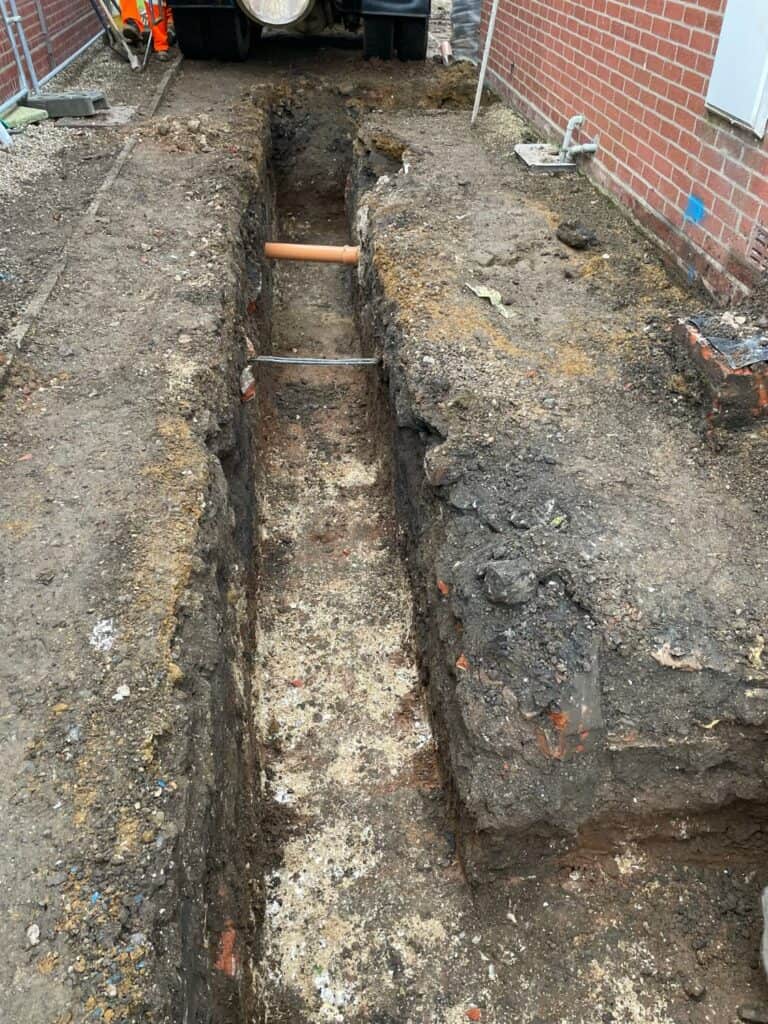 Excavated trench for pipework by brick building.